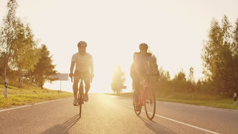 Dos-Ciclistas,-Un-Hombre-Y-Una-Mujer,-Viajan-Por-La-Carretera-En-Bicicletas-De-Carretera-Con-Cascos-Y-Ropa-Deportiva-Al-Atardecer-En-Cámara-Lenta